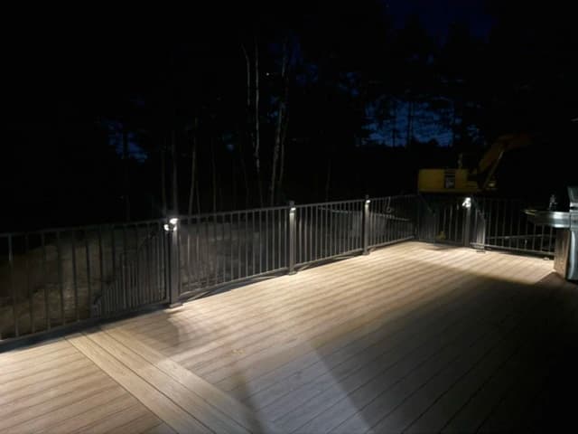 Large composite deck at night with post-cap lighting and black metal railing, wooded backdrop