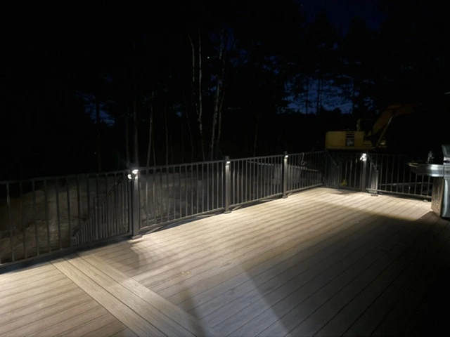 Large composite deck at night with post-cap lighting and black metal railing, wooded backdrop